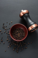 Pepper mill with copper inserts on black background with some black pepper in a clay bowl.