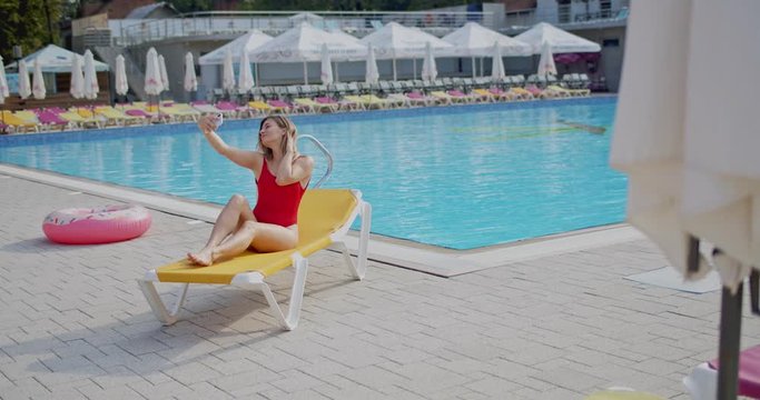 Caucasian young pretty girl in the red swimming suit sitting on the lounge and smiling while taking selfie photos on the smartphone at the pool while resting.