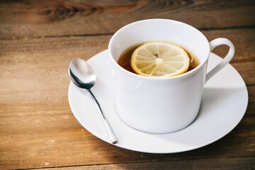 White cup filled with green tea with a slice of lemon on top of a white ceramic plate with a spoon on the side with a wooden background