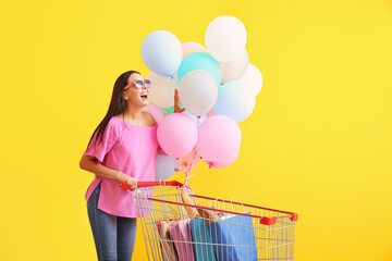Young woman with shopping cart and balloons on color background
