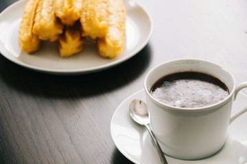 closeup of a white ceramic mug filled with hot chocolate on a ceramic plate with a spoon on the side with a tower of churros on the background
