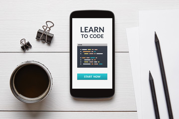 Learn to code concept on smart phone screen with office objects on white wooden table. Top view