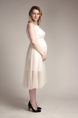 a pregnant woman on a light background in a white dress, holding a large belly, standing sideways in the air