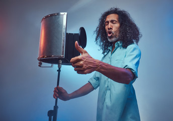African vocal artist sings a song loudly next to a microphone condenser. Isolated on a blue...