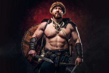 Fototapeta na wymiar Serious Viking clad in light armor with a shield behind his back holds a sword and an axe. Posing on a dark background with red light