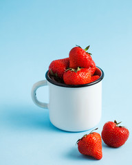 Lots of red ripe fresh strawberries in an enameled white mug on a blue background. There is room for text.
