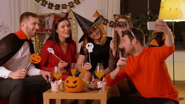 friendship, holiday and people concept - group of happy smiling friends in halloween costumes of vampire, devil, witch and leopard taking selfie by smartphone at home party at night