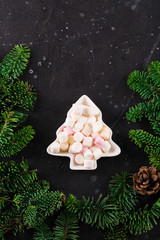 Marshmallows in Christmas tree shape plate on dark background with fir tree green branches. Smores for coffee or hot chocolate, copy space