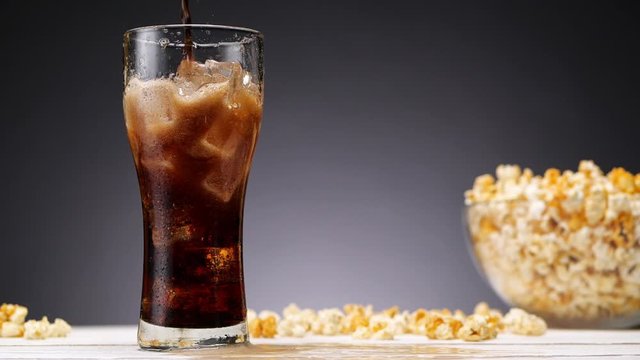 Cola pouring in the glass with ice. Making splashes. Bowl of popcorn on the background. No people. Slow motion.