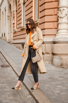 authentic street style portrait of an attractive woman wearing trench coat, sunglasses and slingback shoes, crossing the street.fashion outfit details perfect for autumn fall winter