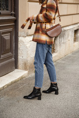 street style portrait of an attractive woman wearing plaid check jacket coat, denim jeans, black ankle boots and brown leather bag, crossing the street. fashion outfit perfect for autumn fall winter