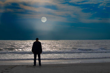 A man stands alone on at the edge of the  calm ocean  looking at rising moon - Powered by Adobe