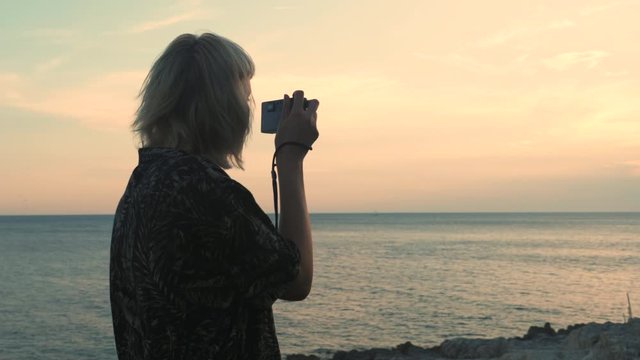 Young blonde woman takes pictures of the sunset sea with an analog camera. She is on summer vacation. Trying to be a photographer.