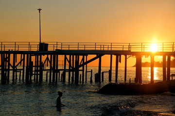 Silhouette of a girl who is swimming in the sea near the pier during sunset.