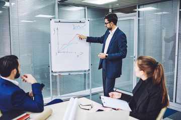 Intelligent male leader of company standing near flip chart during presentation for colleagues,...