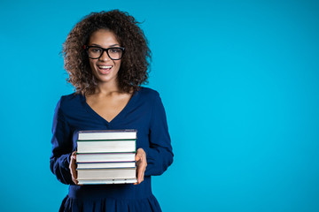 African student with glasses on blue background in the studio holds stack of university books from library. Girl smiles, she is happy to graduate. Copy space.