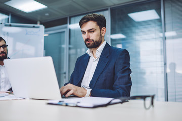 Concentrated male lawyer in formal wear booking tickets on website while using modern laptop device with wireless internet connection in conference room, concept of business people and technology