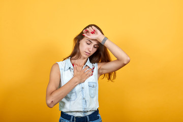 Sick caucasian young woman in blue denim shirt keeping hand on head like temperature, holding other on chest isolated on orange background in studio. People sincere emotions, lifestyle concept.