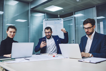 Overjoyed male manager in formal wear feeling excited from received email with good information from business partner while professional colleagues sitting near at desktop and developing project