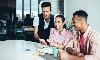 Fototapeta premium Cheerful male and female colleagues reading funny publication in social networks via app on cellphone during break from collaboration on startup, happy group of people smiling from joke on website