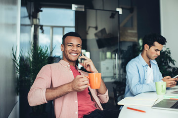 Portrait of cheerful male software developer with coffee cup enjoying communication via application on smartphone gadget during working day with professional colleague, concept of technology