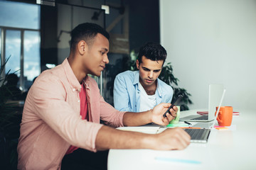 Male diverse employees watching video online via smartphone gadget sitting at desktop with computers, clever colleagues enjoying communication togetherness reading news on web page connected to 4g