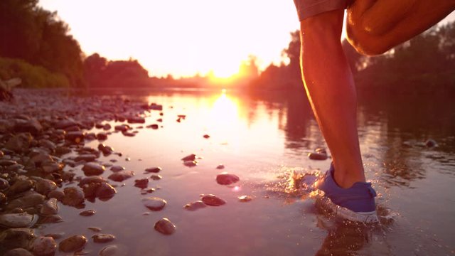 SUPER SLOW MOTION, CLOSE UP Unrecognizable young man running in river shallows at golden sunset. Active sportsman having a morning run in shallow water. Male runner jogging, splashing water at sunrise