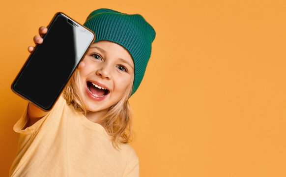 Smiling Screaming Little Girl Kid In Green Modern Winter Hat Showing Blank Screen Of New Popular Mobile Phone Gadget