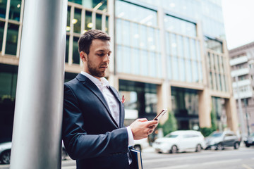 Caucasian male entrepreneur checking mail notification via cell smartphone gadget standing at urban setting in financial district, intelligent businessman using 4g wireless for networking via mobile