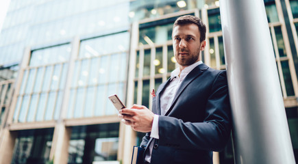 Caucasian intelligent businessman thoughtful looking away while standing at finacial district street and waiting colleague, serious male broker formally dressed using cellphone device outdoors