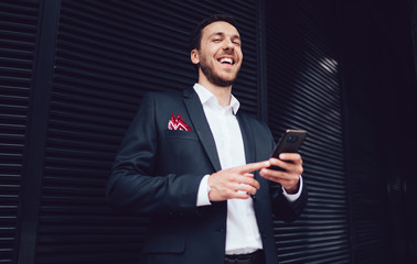 Cheerful man in formal wear standing at urban setting and laughing happy from successful business exchange deal, accomplished Caucasian male proud ceo holding cellphone gadget and smiling outdoors