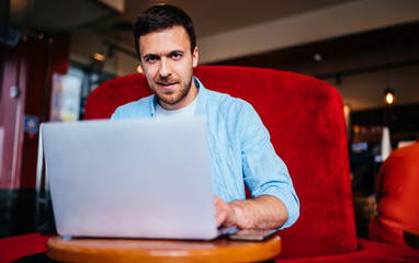 Portrait of skilled male student dressed in casual wear looking at camera during e learning process, handsome man working remotely in coworking space sitting at laptop for online web programming
