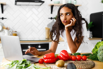 Young african girl has thinkful look typing something in a laptop on a kitchen desk with vegetables