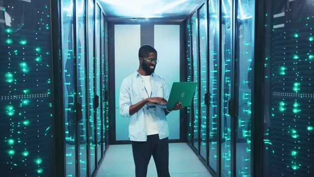 Joyful smiling young IT admin working on laptop computer in modern cyberspace. Positive afro-american specialist inspecting server room of secure data center.