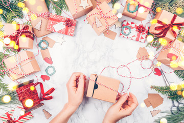 Craft boxes and wraps packed as Christmas gifts and red ribbons on white marble background with festive bokeh lights. The woman packs Christmas gifts. Holiday season concept. Horizontal