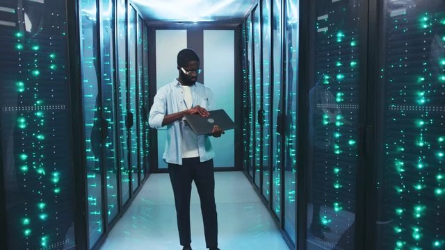 Afro-american young IT specialist working in high secure server storage room. Cheerful employee using laptop computer smiling staying in modern database center.