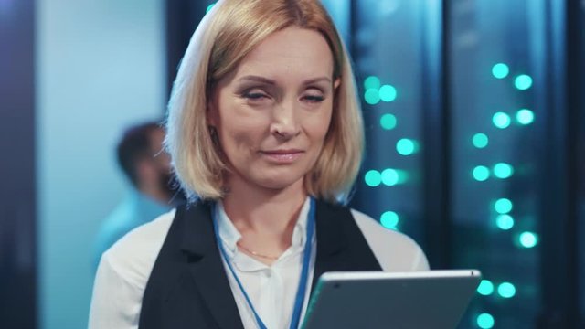 Woman IT server engineer using a tablet working at professional data center. Portrait positive female specialist smiling at camera coworking in high secure server room.