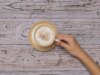 Hand raises Cup with coffee with cream over wooden table.