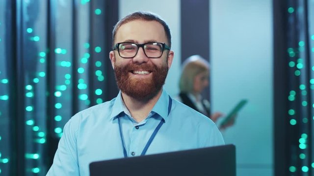 Handsome bearded IT administrator with laptop smiling cheerfully on camera coworking in cyber security cloud computing webhosting at data center.