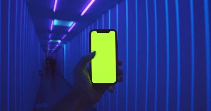 Man hand using green screen smart phone mobile in blue fluorescent corridor interior. Croma-key display mobile. Mock-up. Web design. Online video stream app. Social media. Night club party. Tap, swap.