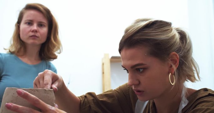 Close up portrait of serious concentrated blonde woman learning to create handmade clay flower pot with teather in craft studio. Beautiful young female hands sculpting. Traveling art business.