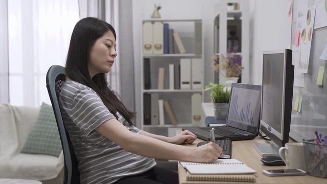 side view of beautiful asian chinese pregnant business woman writing in her notebook while sitting in working place at home. young elegant future motherhood concentrated typing on computer in house.