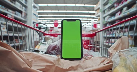 Time-lapse of shopping cart with foodstuff moving along sections and aisles in supermarket. Mock-up smartphone with greenscreen shopping list app inside trolley.
