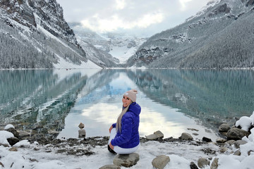Woman sitting on rock looking at scenic view of calm lake and reflections of surrounding mountains with snow, glaciers and trees. Early winter on Lake Louise in Banff National Park. Alberta. Canada
