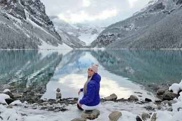Fototapeta premium Woman sitting on rock looking at scenic view of calm lake and reflections of surrounding mountains with snow, glaciers and trees. Early winter on Lake Louise in Banff National Park. Alberta. Canada