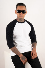 Young man wearing raglan 3/4 sleeve t shirt isolated on white background. Young hipster man with tattoo wearing sunglasses sitting in a chair while posing. Ready for your mock up template design or ba