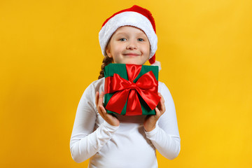 Happy child in santa hat is holding christmas present. Little girl in a white T-shirt on a yellow background