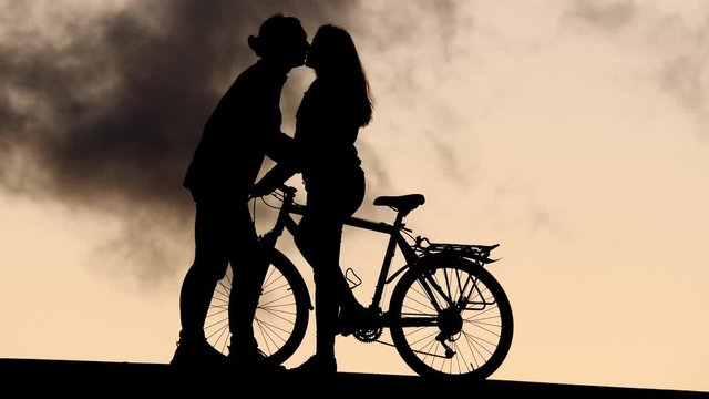 Young woman stand with bike at road side, man come close and give welcome kiss. Silhouetted shot of pair meeting