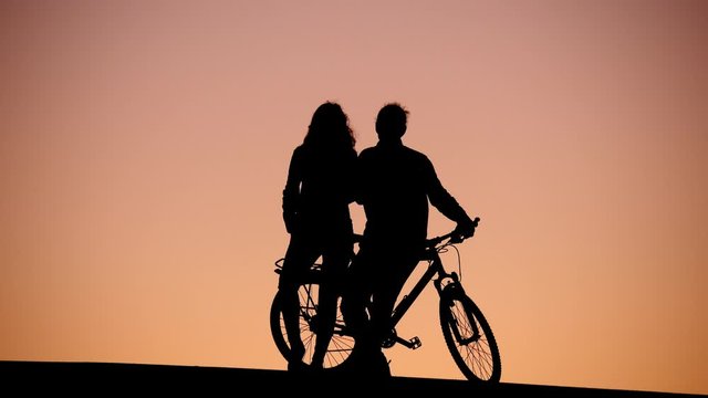 Couple stand at top road, looking to sunset, silhouetted shot against dim colored red sky. Man lean on bicycle, woman stay close