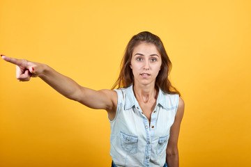 Caucasian young woman in blue denim shirt pointing into distance, looking confused isolated on orange background in studio. People sincere emotions, lifestyle concept.